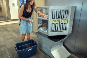 A student wearing a backpack helps restock a gray food pantry from a blue plastic tub.
