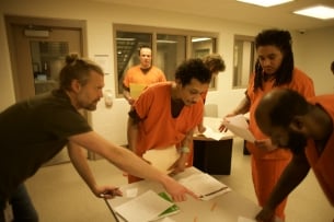 Nate Johnson, founder of FreeWriters, and three incarcerated students in orange uniforms stand around a table covered in papers.