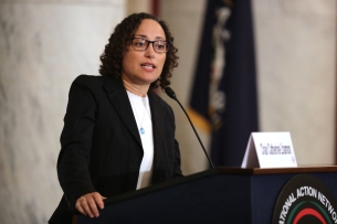 A woman with curly hair wearing glasses and a blazer speaks at a podium