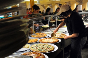A student dining hall worker slices a pizza as other students stand in the cafeteria line