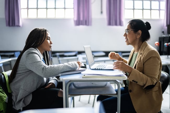 Female professor sits at a desk in an otherwise empty classroom with a young female student, talking and gesturing with her hands. Student is listening.