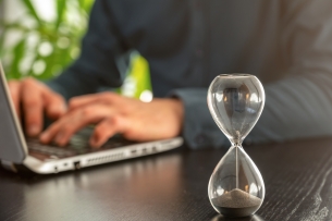 A close-up of an hourglass sitting on a desk; behind the hourglass a person's hands rest on a laptop.