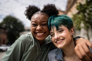 Two young women smile and hug on campus, outside. They're looking at the camera and appear happy.
