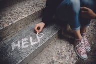 Cropped photo of a student writing "help" in chalk on a set of steps while sitting. Face is not visible.