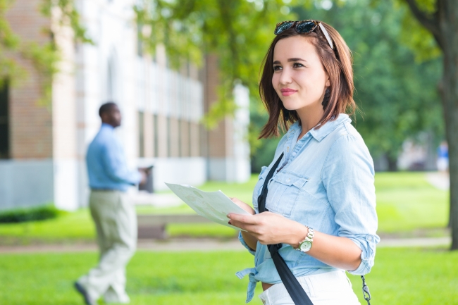 Young woman, presumably on a college tour, smiles as she holds a map in a campus green space.
