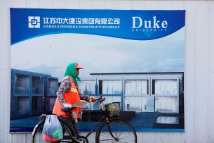 A man wearing a bright orange vest and green hoodie walks his bicycle past a sign for Duke University's campus in Kunshan, China.