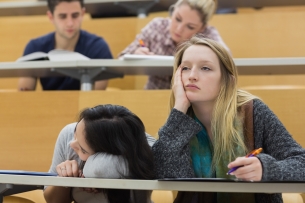A group of disengaged, bored-looking students—one resting her head on her hand, and another with their head on the desk—in a university lecture hall.