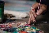 A hand with a paintbrush works on a colorful abstract painting on a wooden table.
