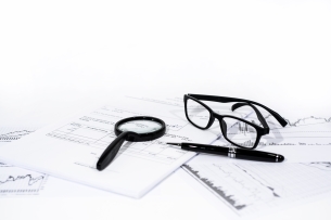 Black magnifying glass, eyeglasses and pen sit on papers with charts and graphs