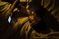 A young Black adult scrolls on his phone while lying down in bed.
