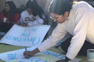 A student writes on a protest sign. Behind her, a student writes on a sign that says "opportunity for all" in blue letters.