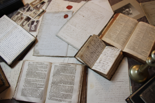A collection of open books and notebooks, plus some black-and-white pictures, strewn on a table, as if being actively used for research. The books have an appearance of oldness. 
