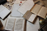 A collection of open books and notebooks, plus some black-and-white pictures, strewn on a table, as if being actively used for research. The books have an appearance of oldness. 