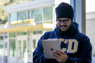 A male student wearing a UC Davis sweatshirt stands by a window in the daytime, looking at his e-reader and presumably reading digitally delivered course materials. 
